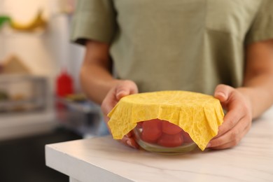 Woman holding bowl of fresh tomatoes covered with beeswax food wrap at table in kitchen, closeup. Space for text