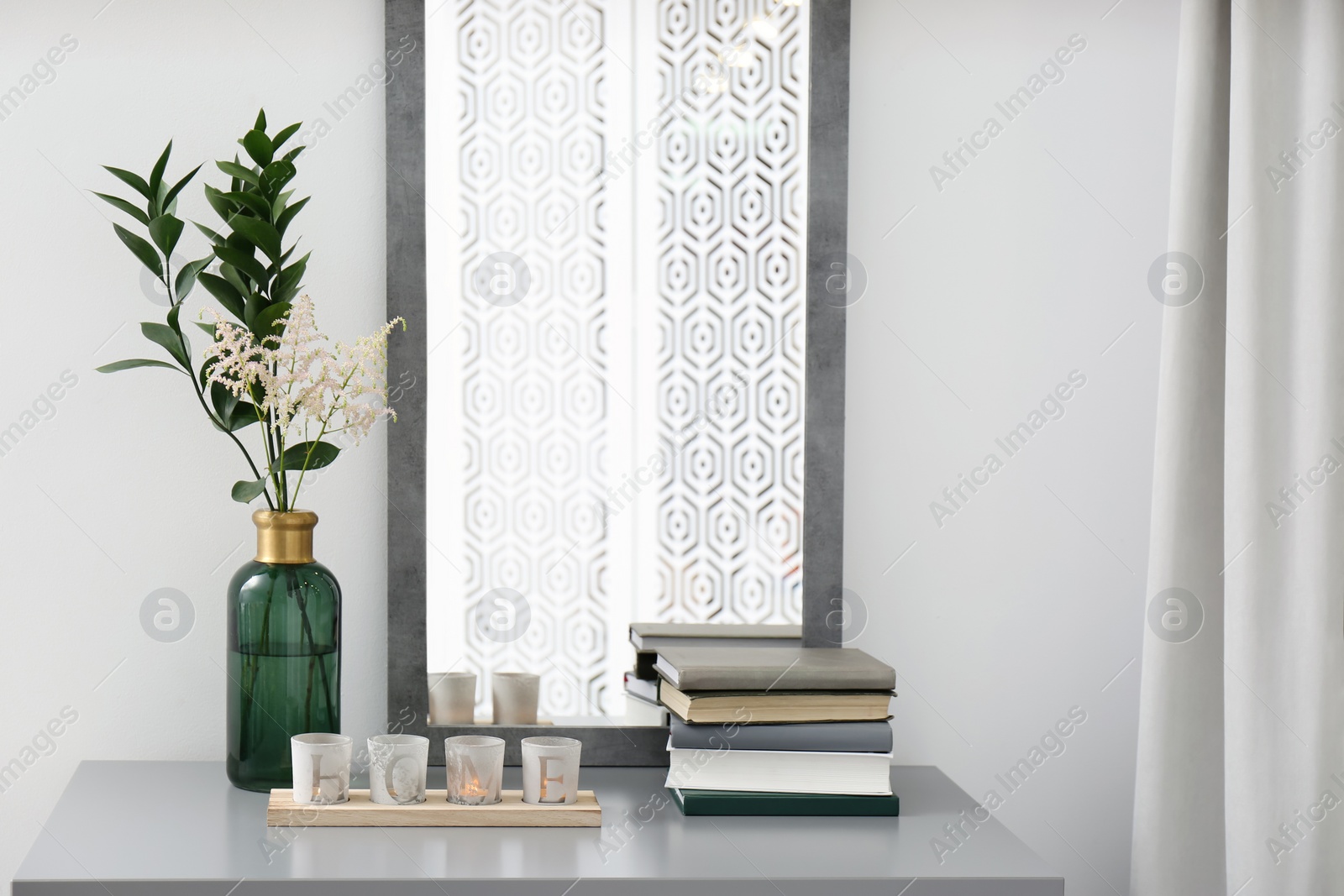 Photo of Vase with beautiful flowers, burning candles in holders and books on grey table indoors. Interior elements