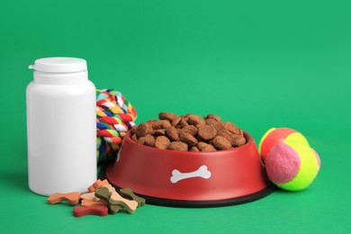 Bowl with dry pet food, bottle of vitamins and toys on green background