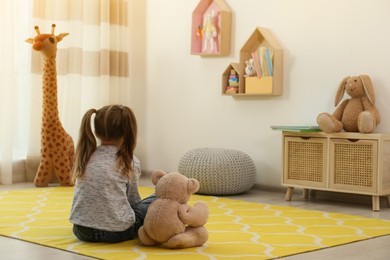 Photo of Lonely little girl with teddy bear sitting on floor at home, back view. Autism concept