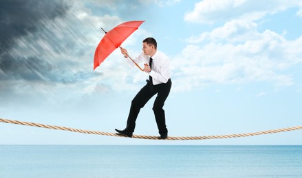 Image of Risks and challenges of entrepreneurship. Businessman with umbrella protecting from coming rainstorm on rope over sea