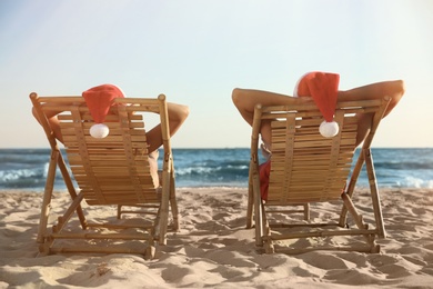 Photo of Lovely couple with Santa hats relaxing on deck chairs at beach, back view. Christmas vacation