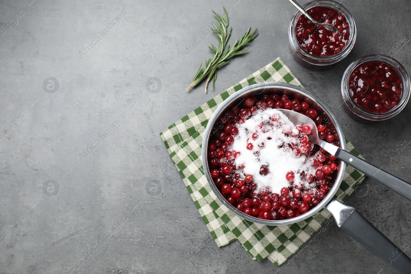 Photo of Fresh cranberries with sugar in saucepan and cranberry sauce in glass jars served on gray table, flat lay. Space for text
