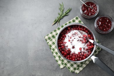 Photo of Fresh cranberries with sugar in saucepan and cranberry sauce in glass jars served on gray table, flat lay. Space for text