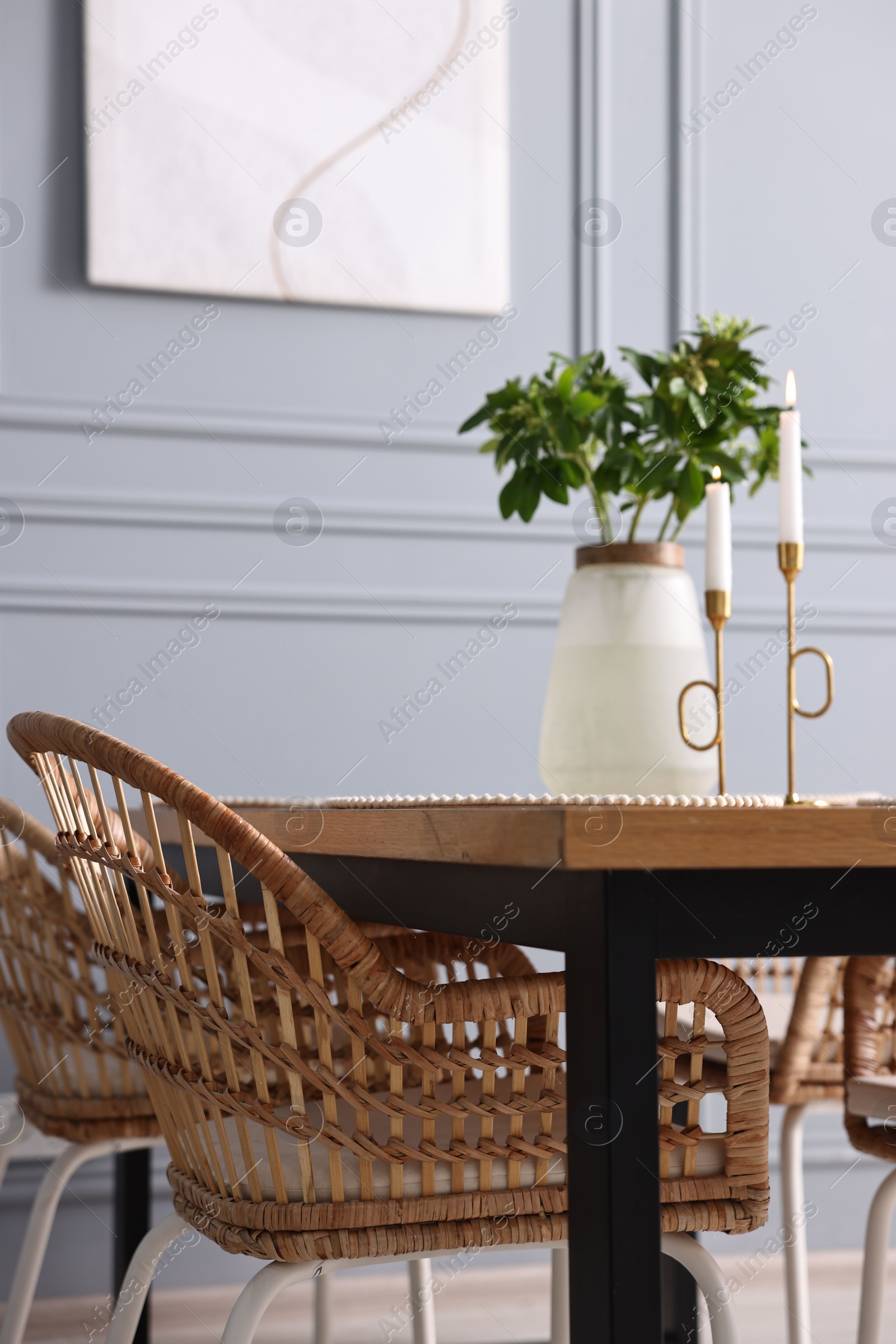 Photo of Rattan chairs, table, burning candles and vase with green branches in stylish dining room