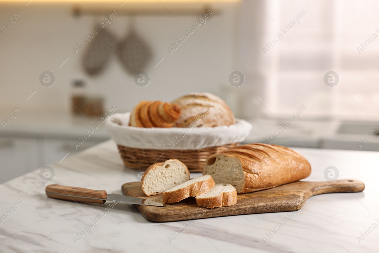 Photo of Wicker bread basket with freshly baked loaves and knife on white marble table in kitchen