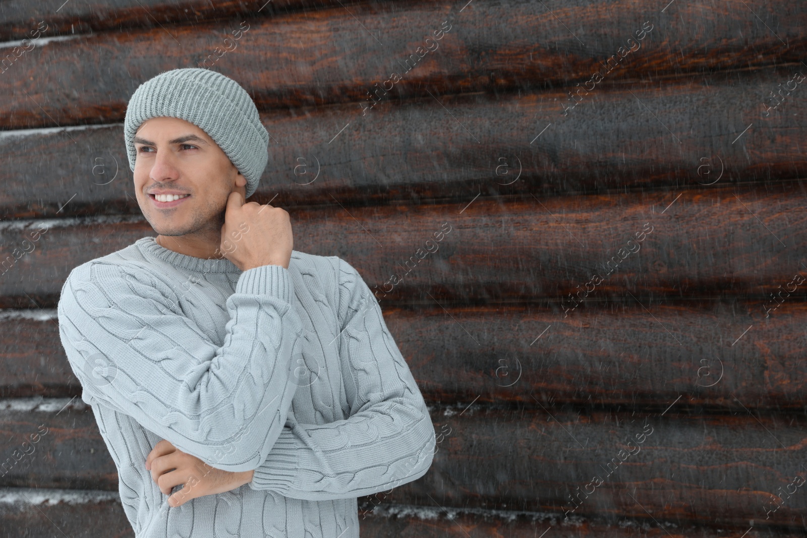 Photo of Handsome man wearing warm sweater and hat near wooden wall, space for text. Winter season