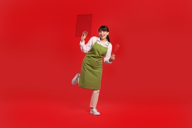 Photo of Happy confectioner in apron holding professional whisk and cooling rack on red background