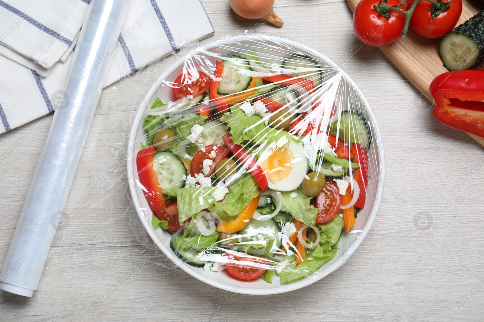 Photo of Bowl of fresh salad with plastic food wrap on white wooden table, flat lay