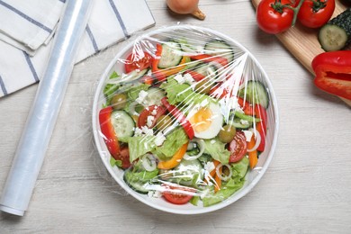 Bowl of fresh salad with plastic food wrap on white wooden table, flat lay