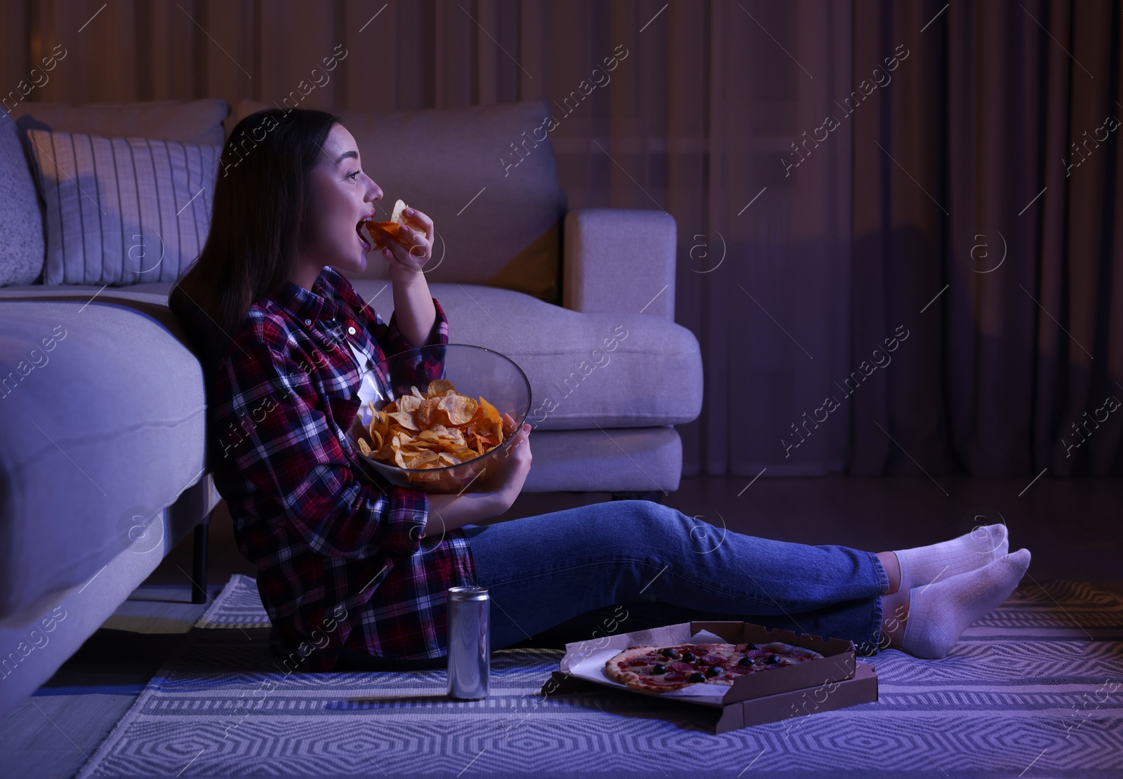 Photo of Young woman eating chips while watching TV in room at night. Bad habit