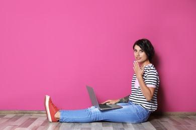 Young woman with modern laptop sitting on floor near color wall