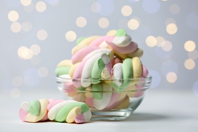 Photo of Glass bowl with colorful marshmallows on white table against blurred lights, closeup