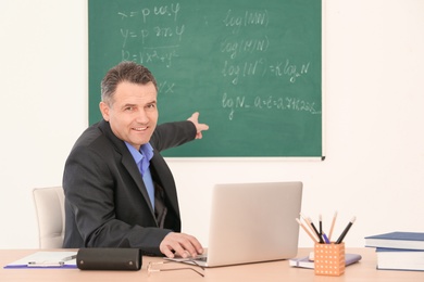 Photo of Male teacher working with laptop at table in classroom