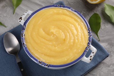 Delicious lemon curd in bowl, green leaves and spoon on grey table, closeup