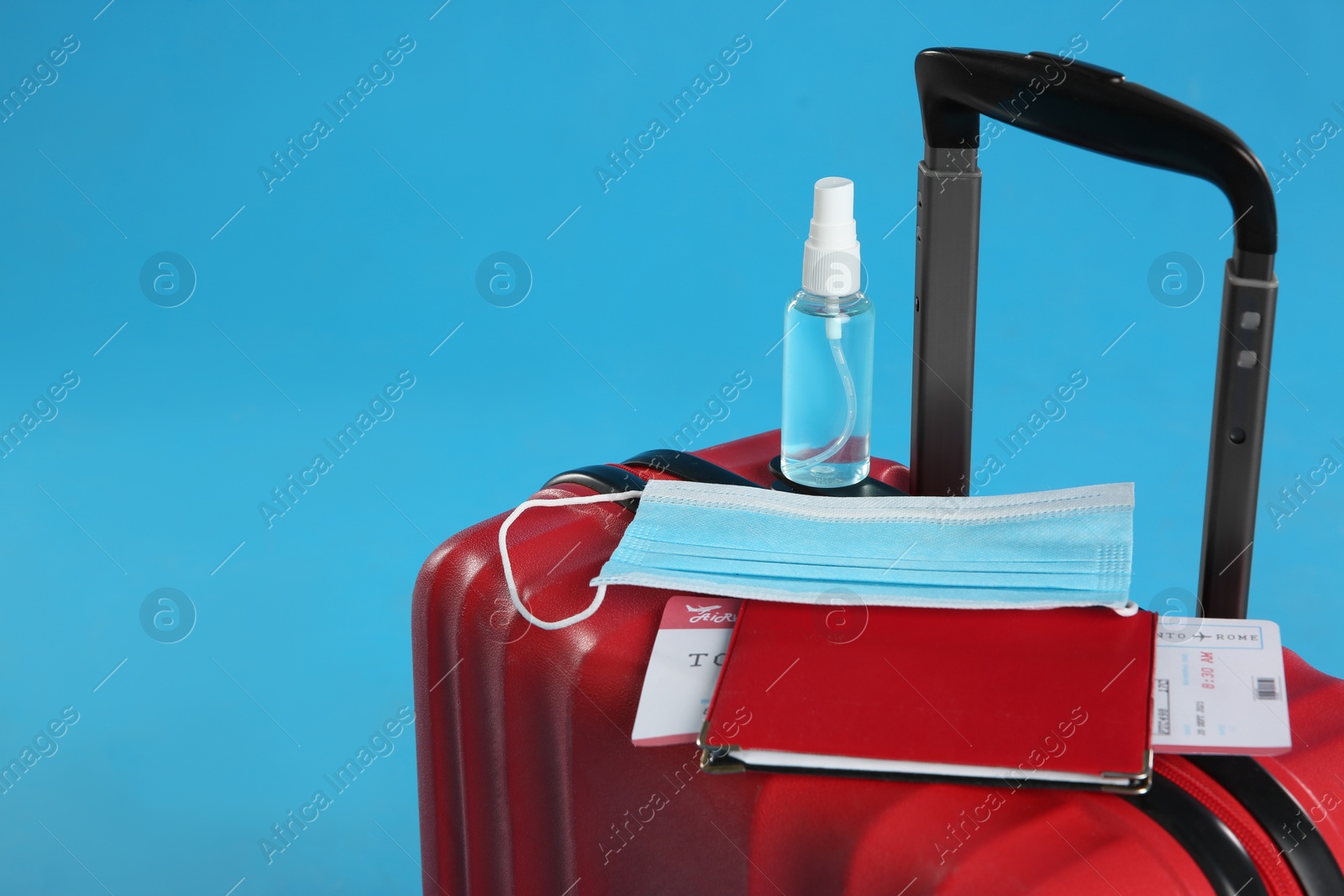 Photo of Stylish red suitcase with protective mask, antiseptic spray, passport and ticket on light blue background, space for text. Travelling during coronavirus pandemic