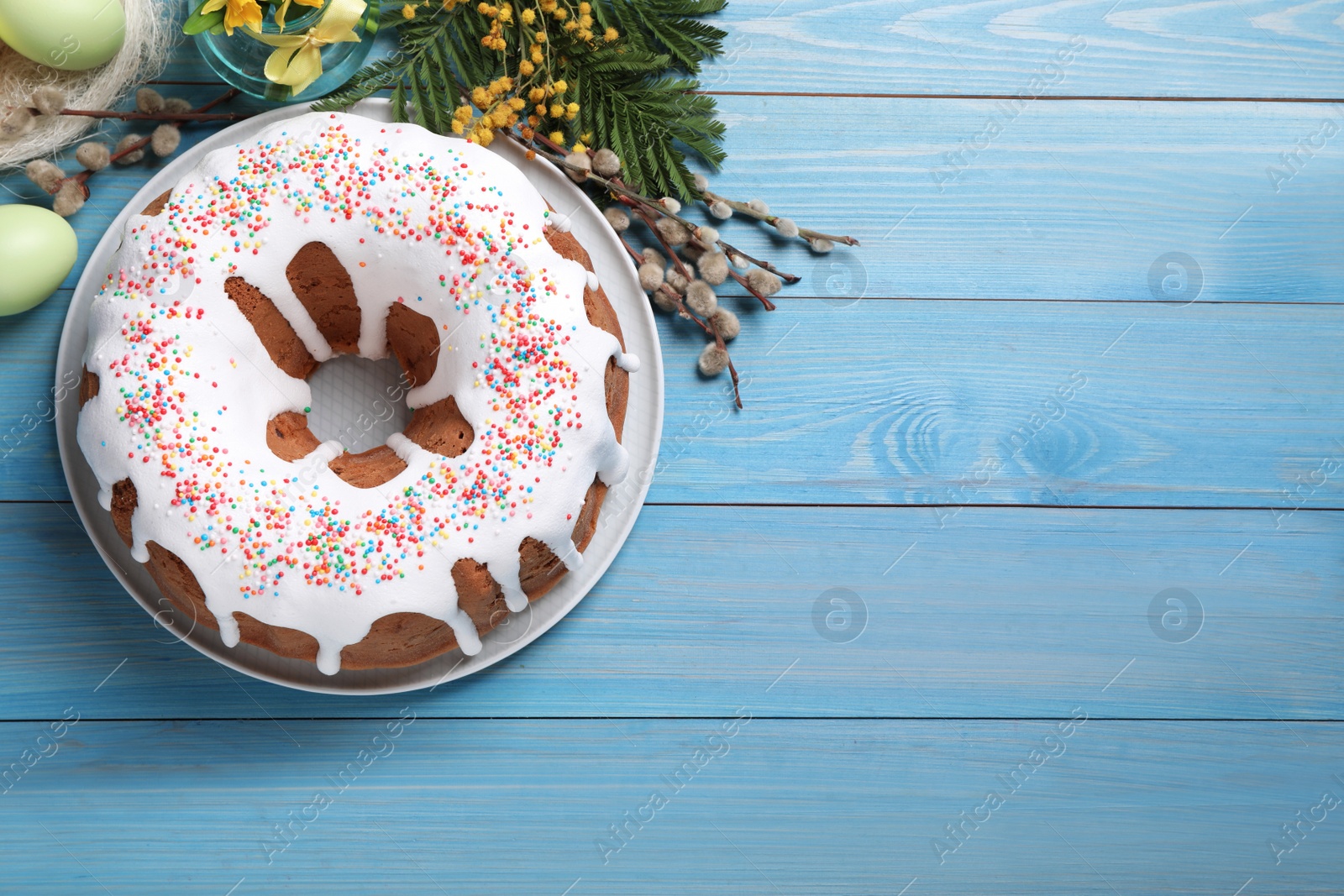 Photo of Glazed Easter cake with sprinkles, painted eggs and flowers on light blue wooden table, flat lay. Space for text