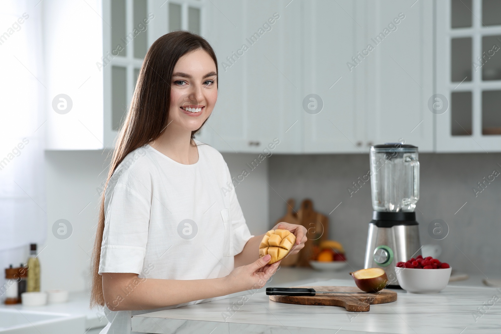 Photo of Beautiful young woman preparing mango for tasty smoothie at white marble table in kitchen