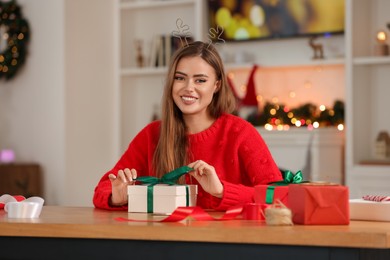 Beautiful young woman in deer headband decorating Christmas gift at table in room