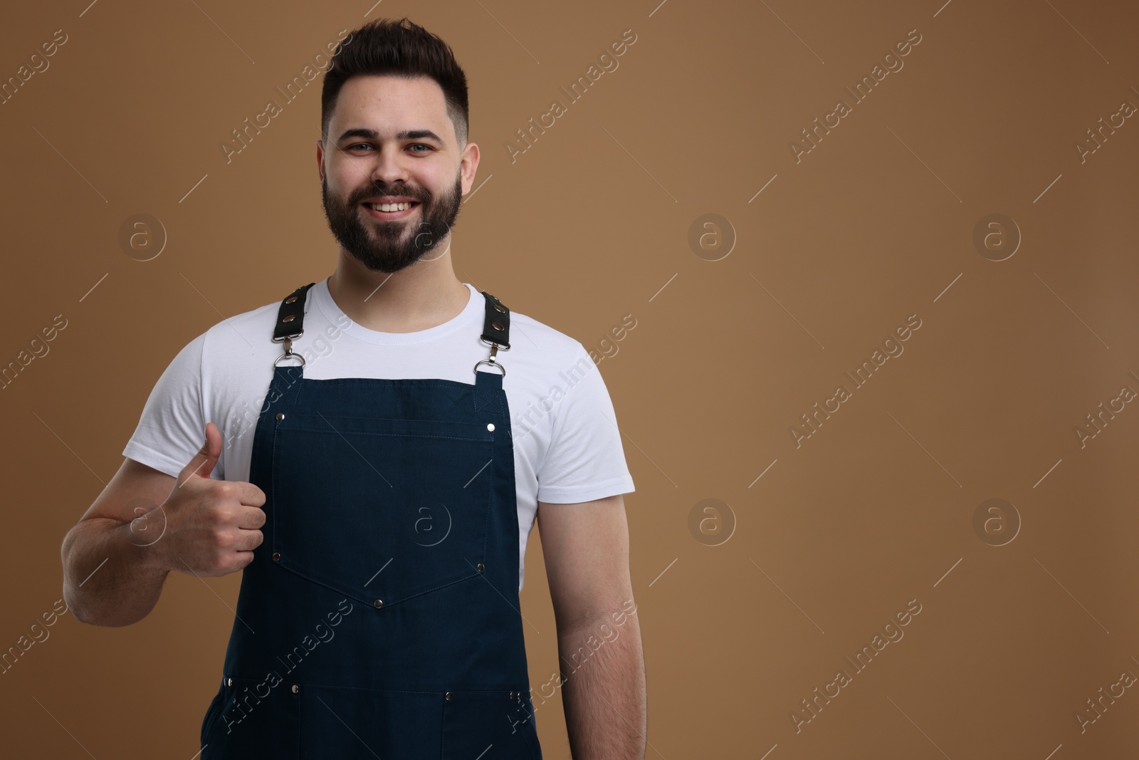 Photo of Smiling man in kitchen apron showing thumb up on brown background. Mockup for design