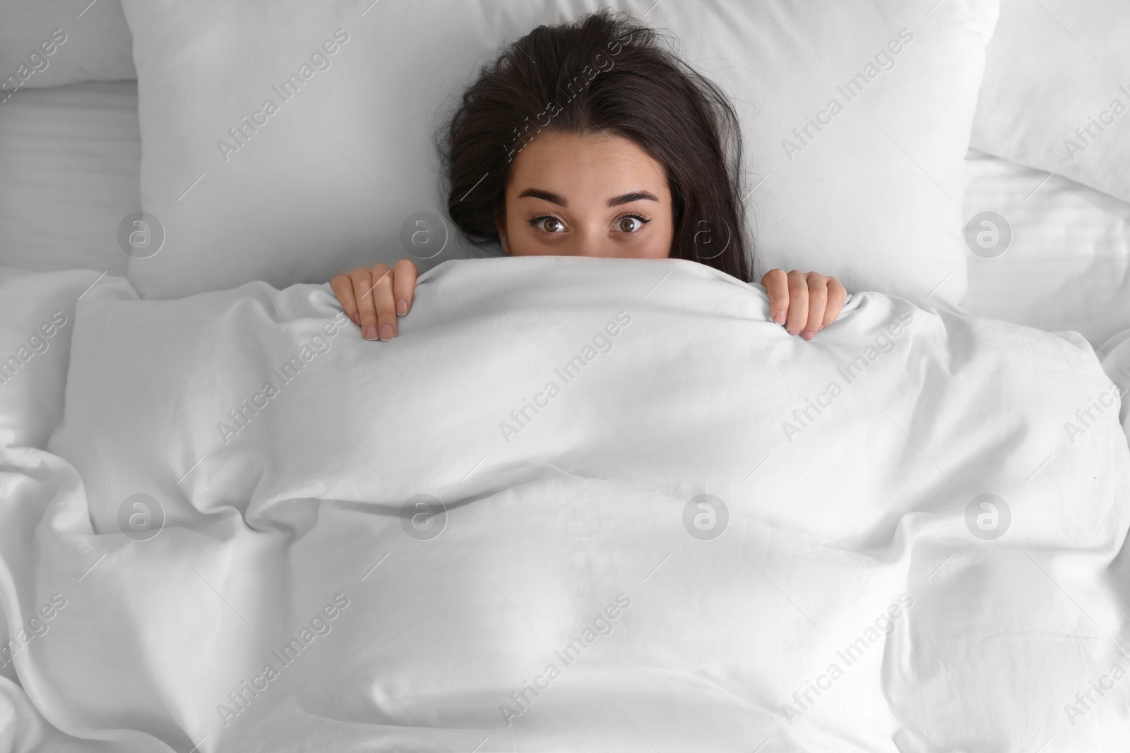 Photo of Young woman hiding under warm white blanket in bed, top view