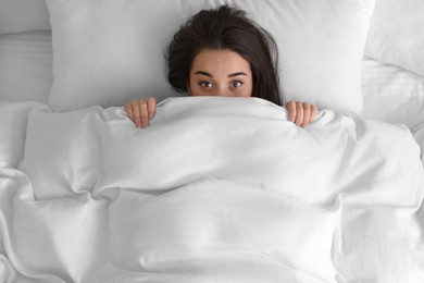 Photo of Young woman hiding under warm white blanket in bed, top view