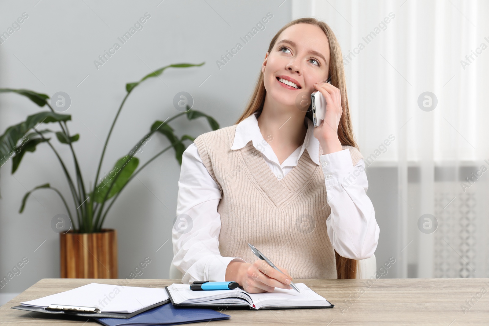 Photo of Woman taking notes while talking on smartphone at wooden table in office