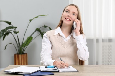 Photo of Woman taking notes while talking on smartphone at wooden table in office