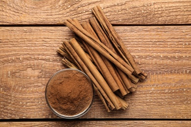 Photo of Aromatic cinnamon powder in bowl near sticks on wooden table, flat lay