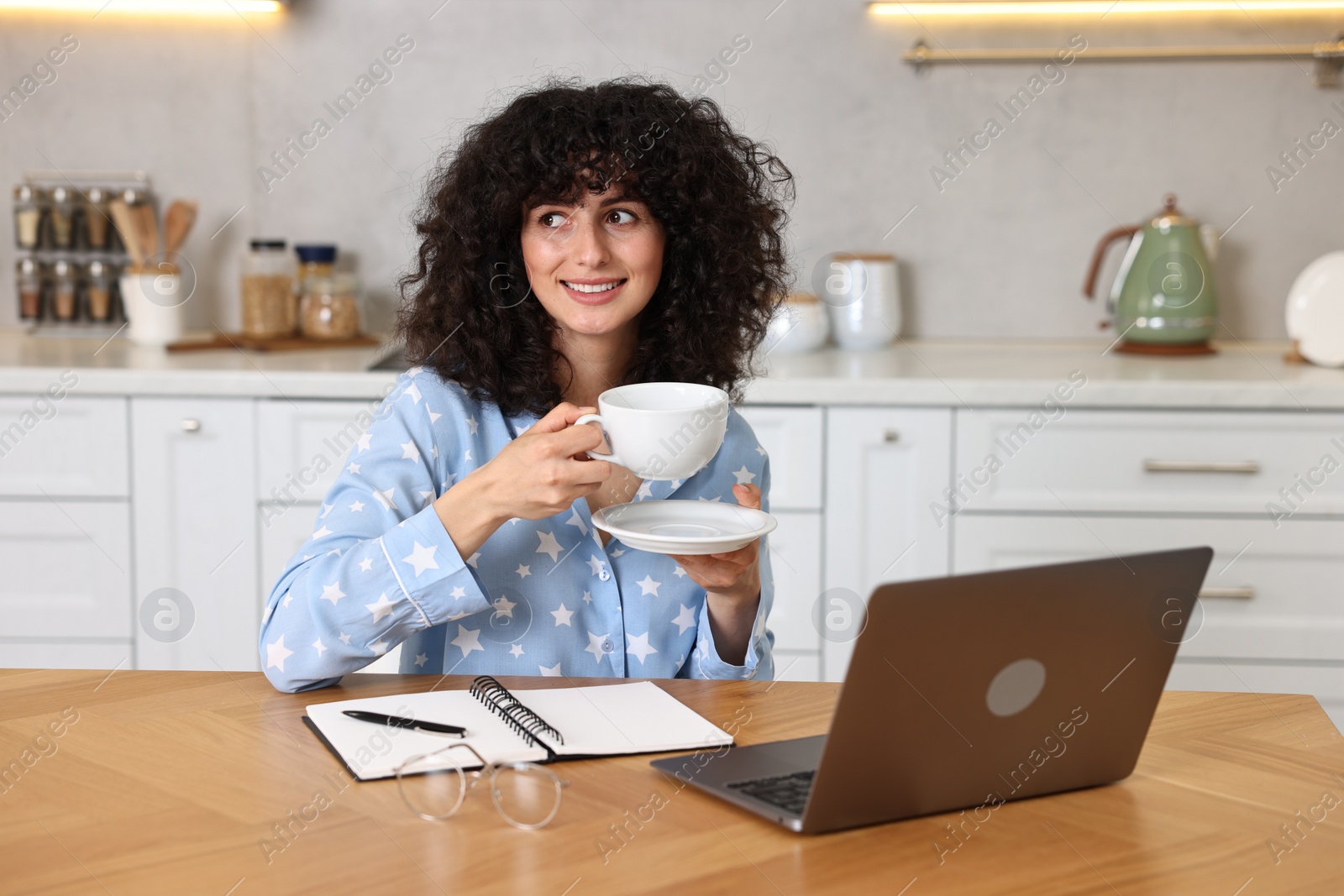 Photo of Beautiful young woman in stylish pyjama with cup of drink at wooden table in kitchen