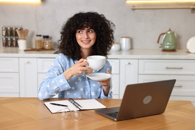 Beautiful young woman in stylish pyjama with cup of drink at wooden table in kitchen