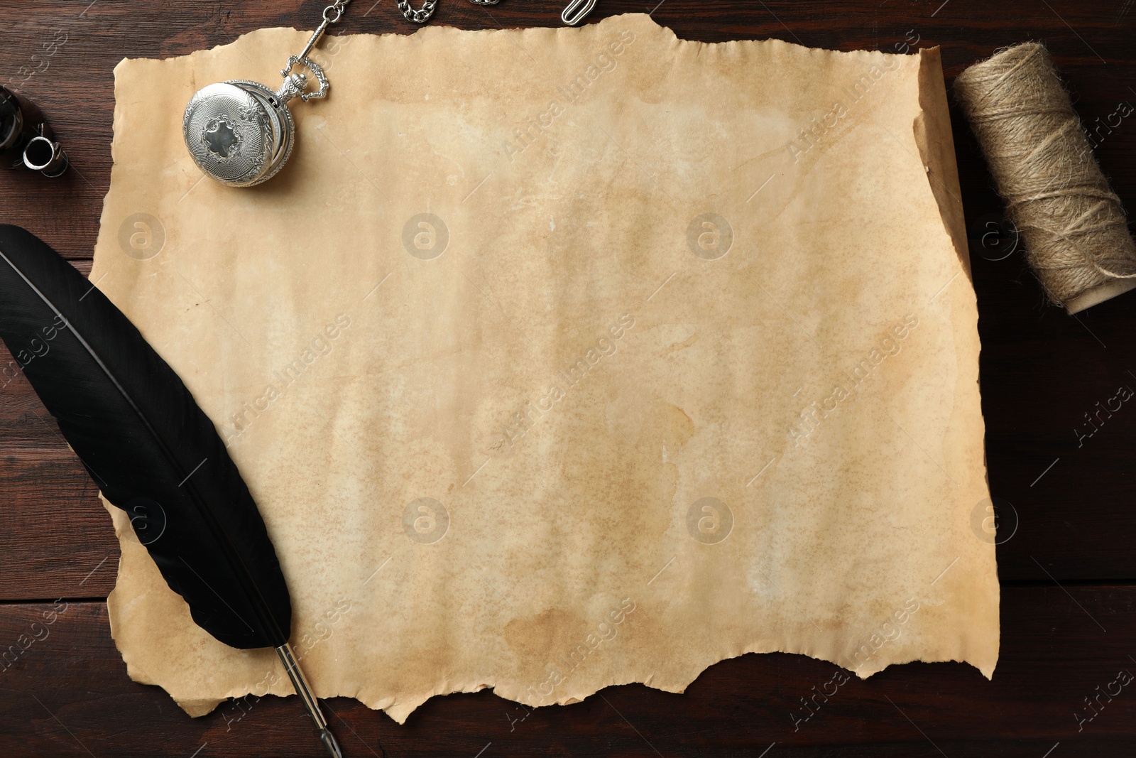 Photo of Sheet of old parchment paper, black feather, inkwell, rope and pocket chain clock on wooden table, flat lay