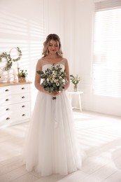 Happy bride wearing beautiful wedding dress with bouquet at home