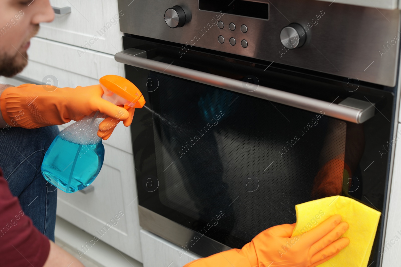 Photo of Young man cleaning oven with rag and detergent in kitchen, closeup