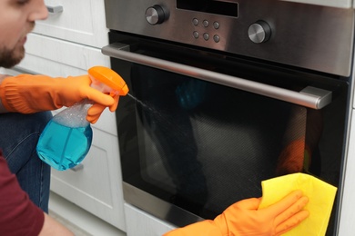 Young man cleaning oven with rag and detergent in kitchen, closeup