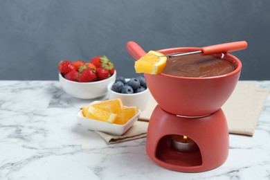 Photo of Fondue pot with melted chocolate, fresh orange, different berries and fork on white marble table. Space for text