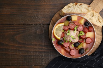 Photo of Meat solyanka soup with thin dry smoked sausages in bowl and bread slices on wooden table, flat lay