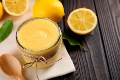 Photo of Delicious lemon curd in glass jar, fresh citrus fruits, green leaves and spoon on wooden table, closeup