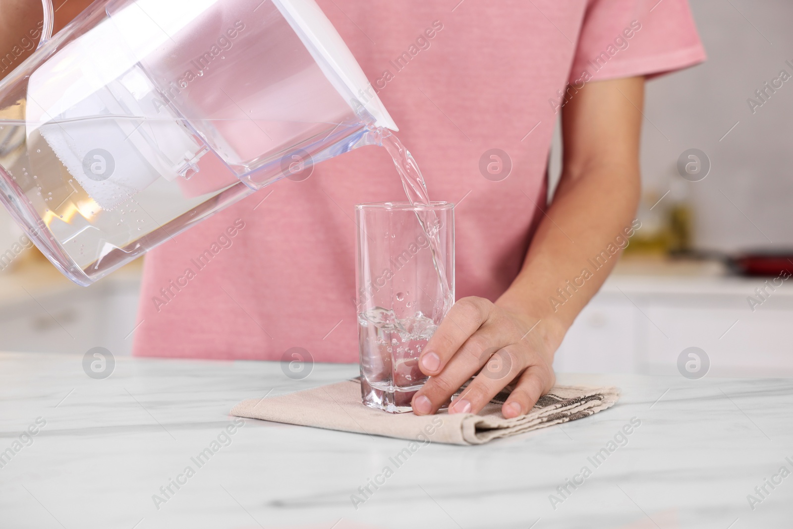 Photo of Man pouring water from filter jug into glass at white marble table in kitchen, closeup