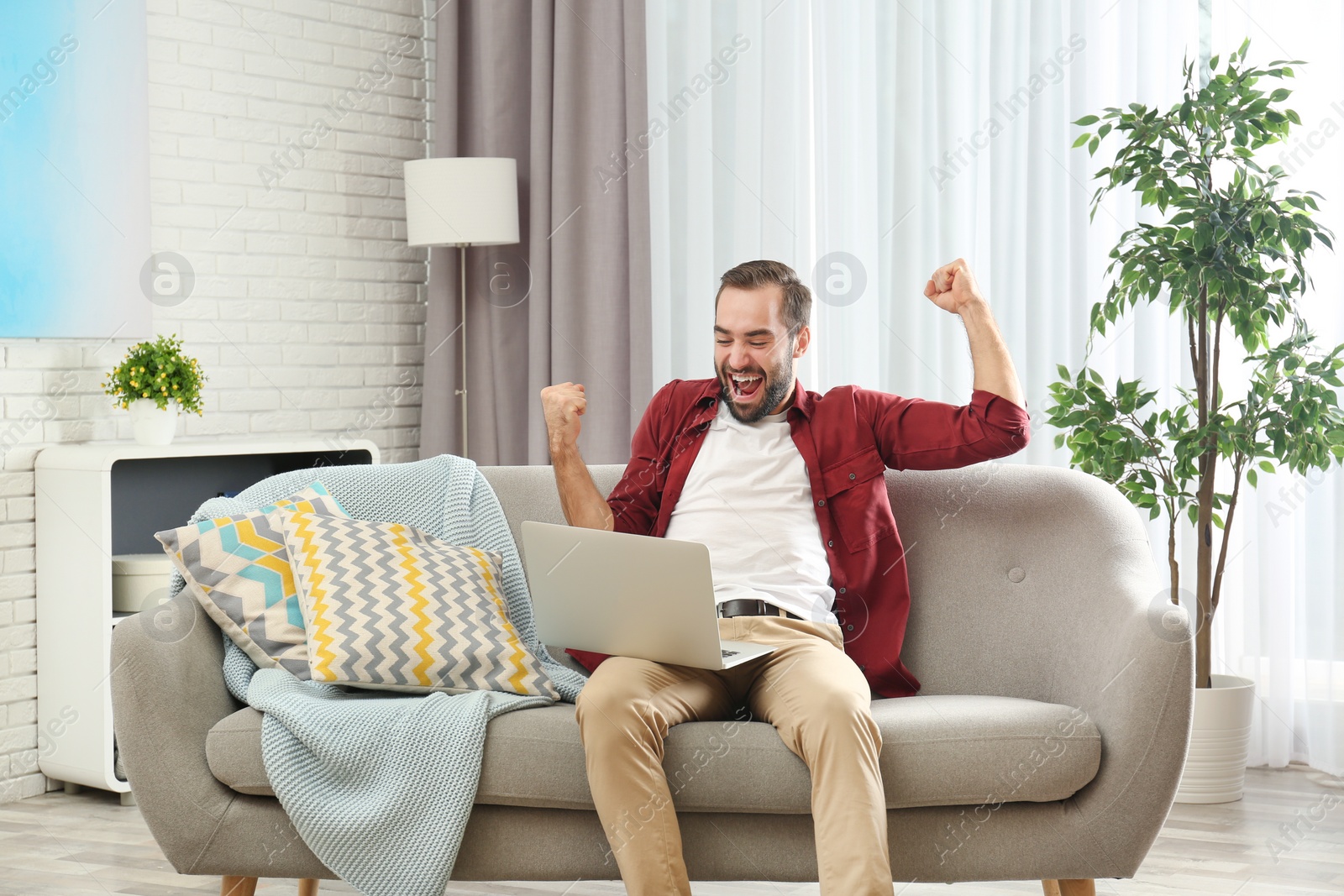 Photo of Emotional young man with laptop celebrating victory on sofa at home