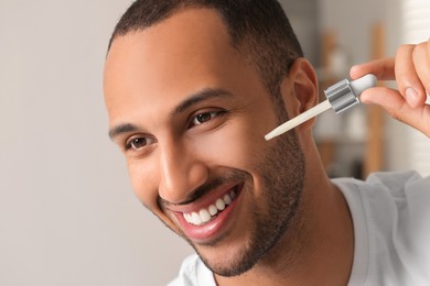 Handsome man applying cosmetic serum onto face indoors, closeup