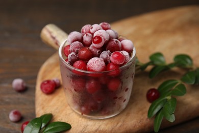 Photo of Frozen red cranberries in glass pot and green leaves on wooden table, closeup