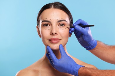 Doctor drawing marks on woman's face for cosmetic surgery operation against light blue background