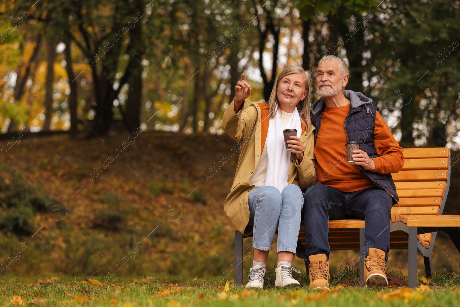 Photo of Affectionate senior couple with cups of coffee on wooden bench in autumn park, space for text