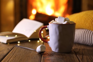 Mug with hot cocoa, marshmallows, lights and book on wooden table near fireplace