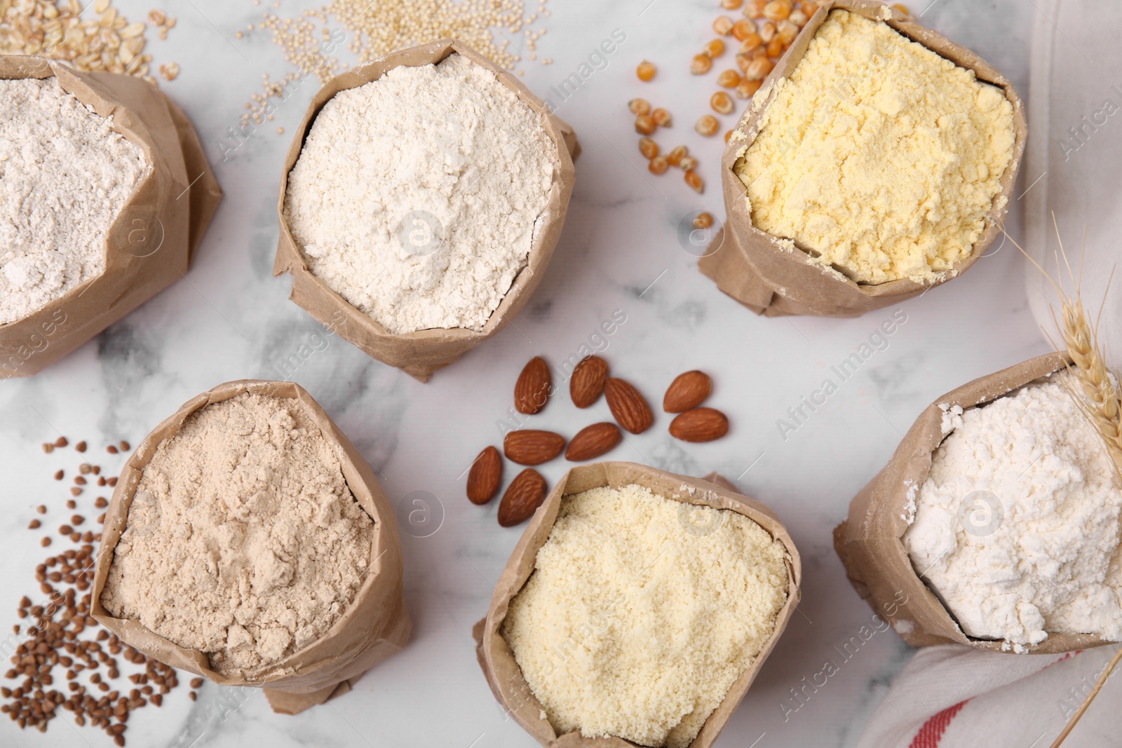 Photo of Paper bags with different types of flour and ingredients on white marble table, flat lay