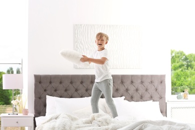 Cute little boy with pillow jumping on bed at home