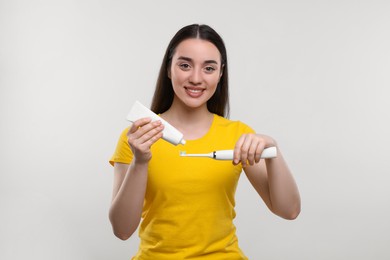 Happy young woman squeezing toothpaste from tube onto electric toothbrush on white background