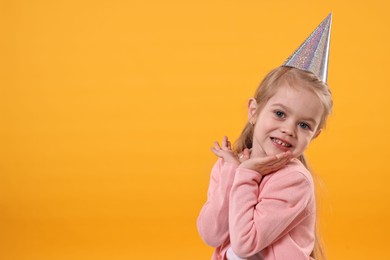 Photo of Birthday celebration. Cute little girl in party hat on orange background, space for text