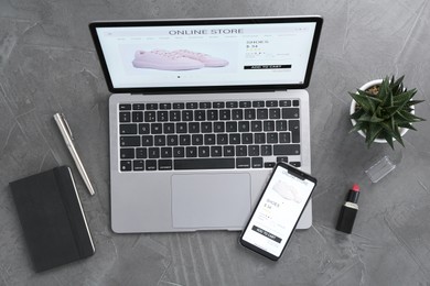 Online store website on laptop screen. Computer, smartphone, stationery, lipstick and houseplant on grey table, flat lay